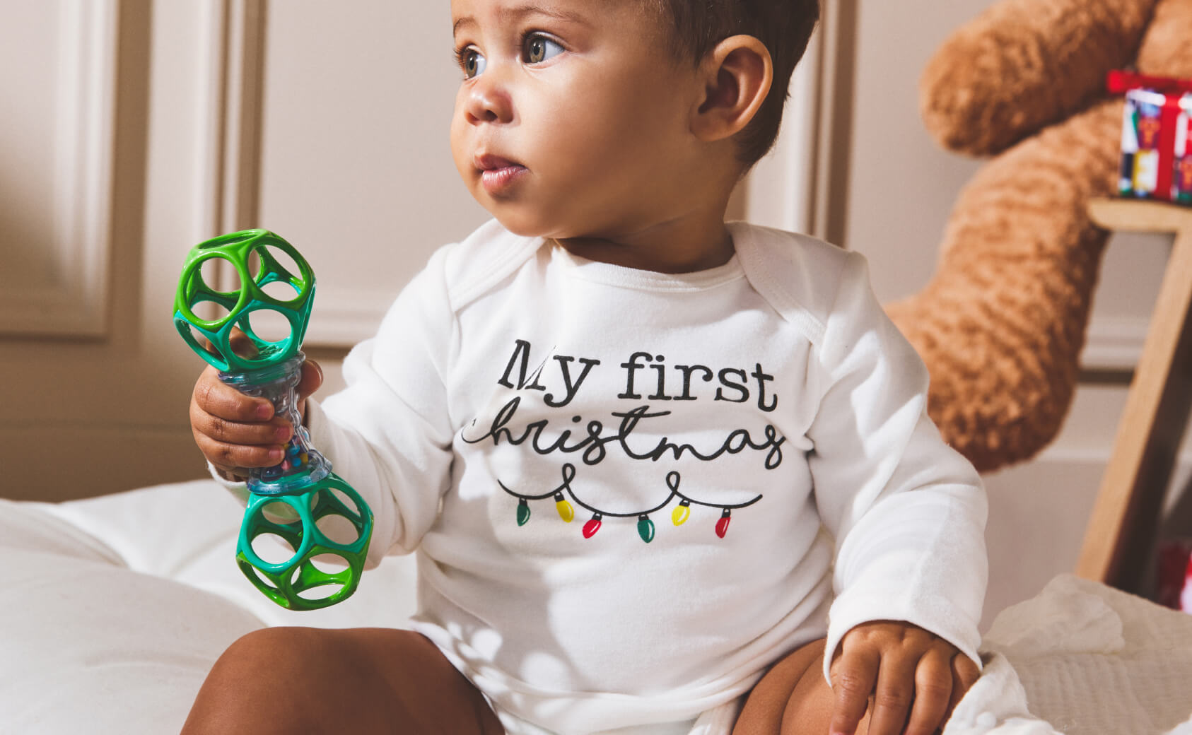 A baby holding a green rattle and wearing a white onesie that says "My First Christmas".