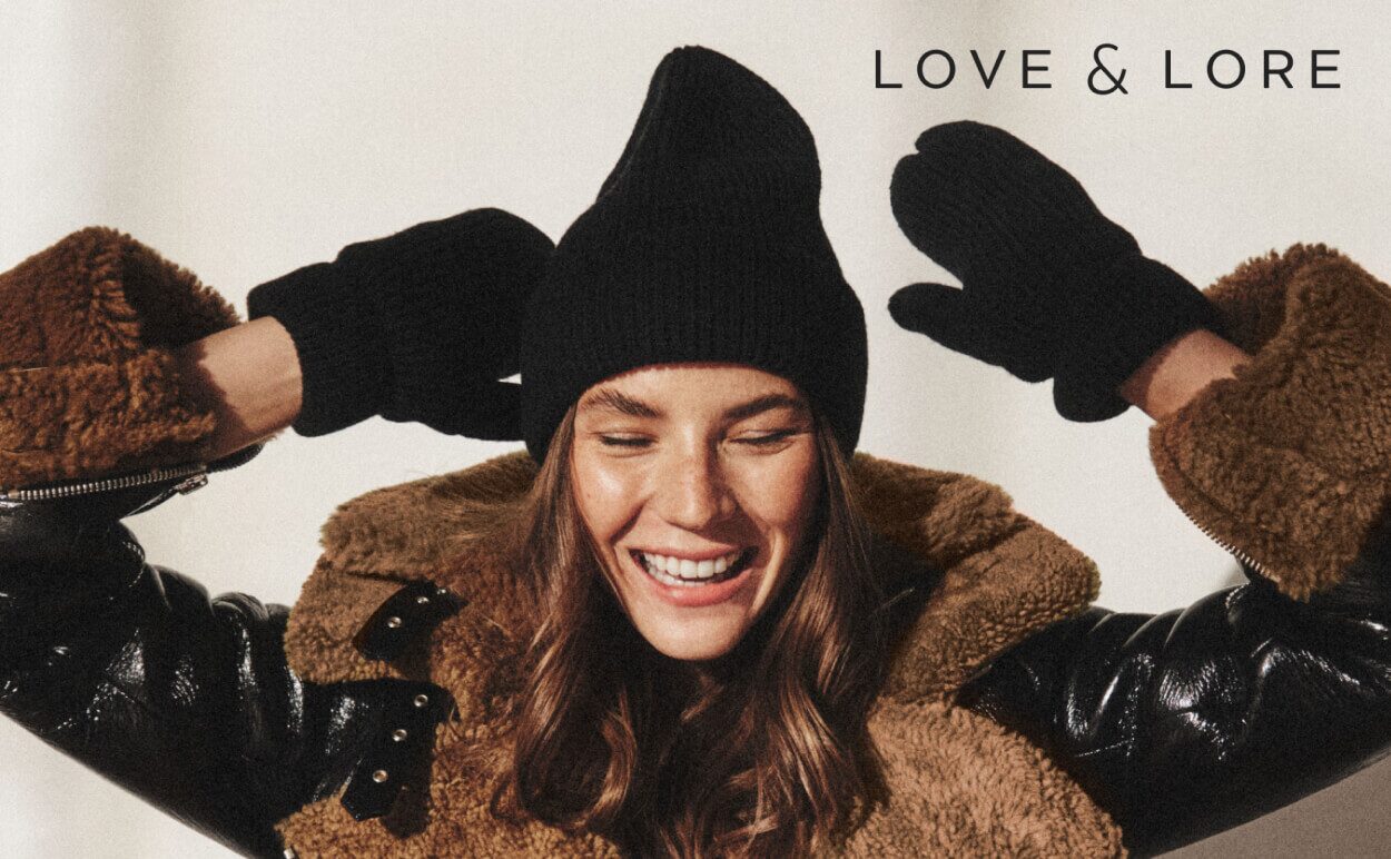A woman smiling while wearing a black Love & Lore beanie and black mittens.