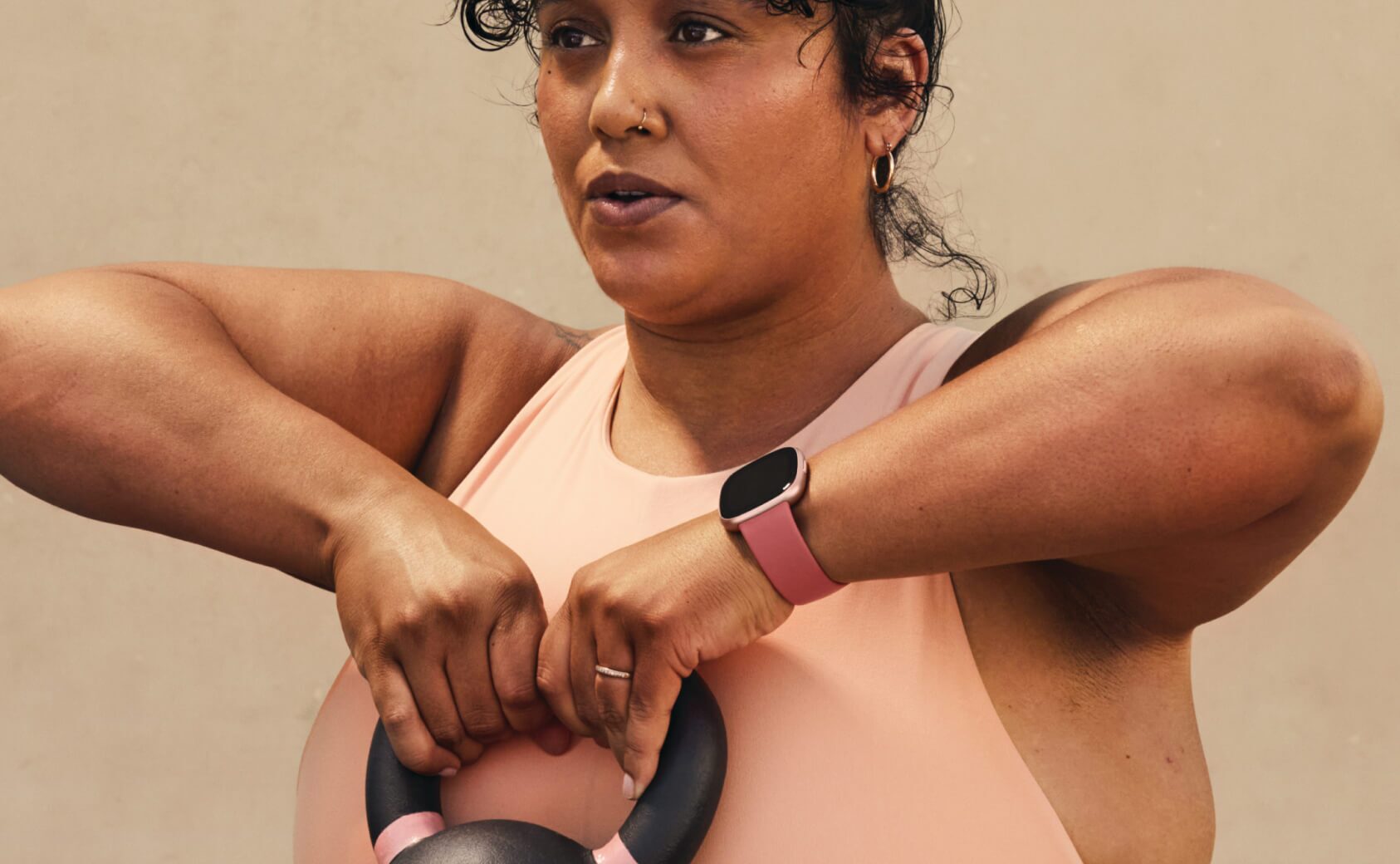 Model lifting a kettlebell wearing a Fitbit Versa 4 smartwatch
