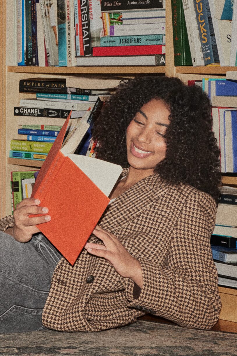 Smiling woman leaning on a bookshelf reading a book.