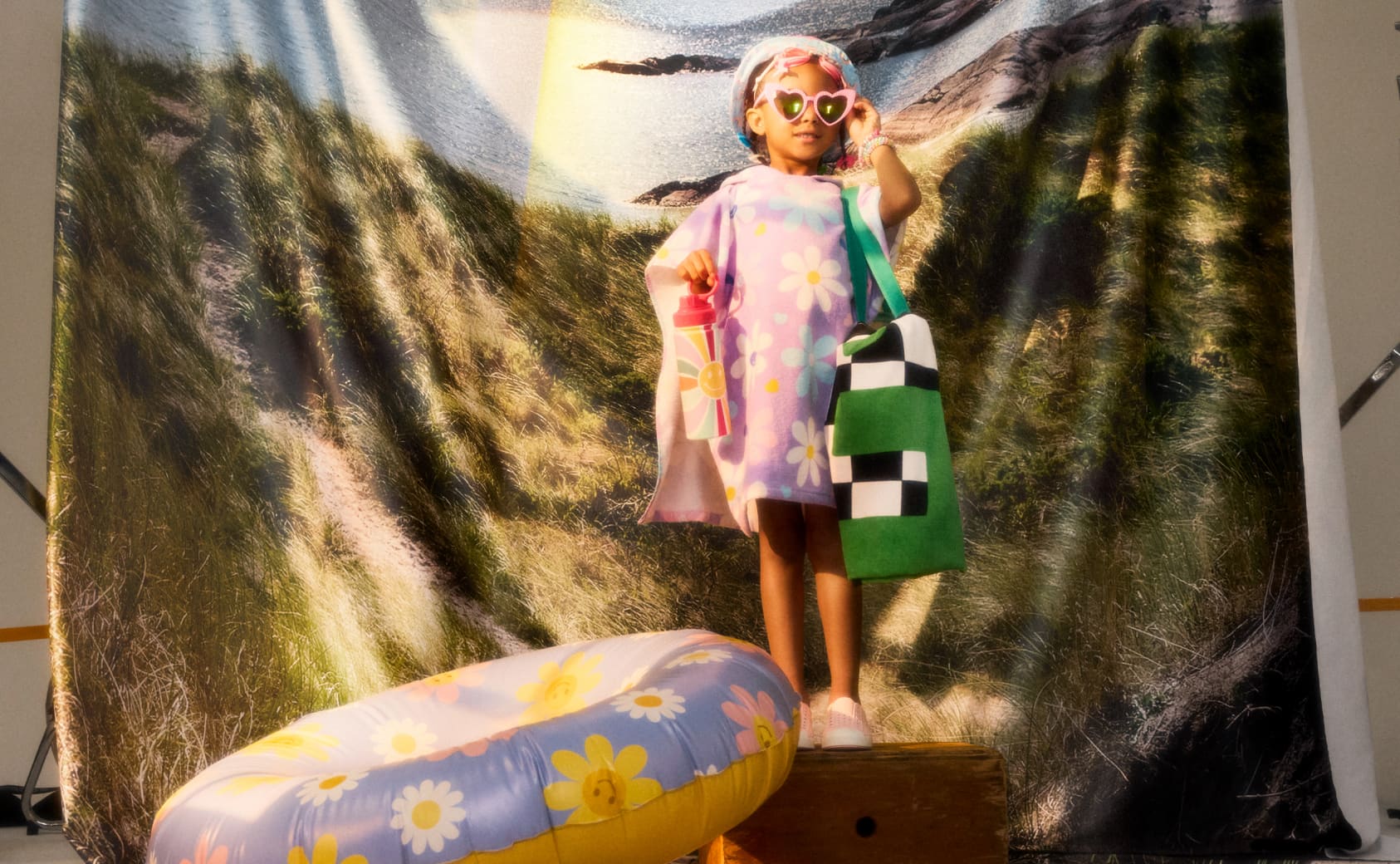 A kid in a towel poncho holding a towel tote and water bottle beside a pool floaty.
