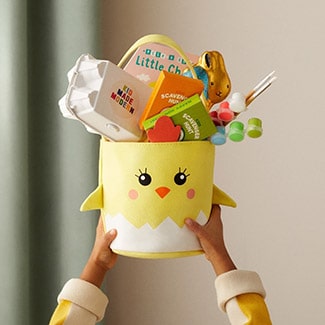 @indigo instagram post: Hands holding up a yellow chick-shaped Easter basket filled with gifts, including a Kid Made Modern craft kit, paints and paintbrushes, two activity books, a chocolate bunny, and the book Peek-A-Boo Little Chick by Yu-Hsuan Huang.